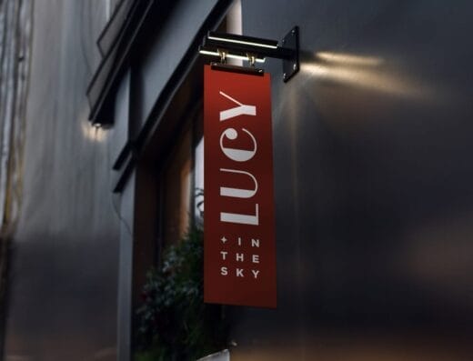 Un panneau vertical rouge comportant le texte blanc en gras « LUCY + IN THE SKY » se dresse bien en évidence sur un mur gris, son message brille de mille feux sous l'éclairage.