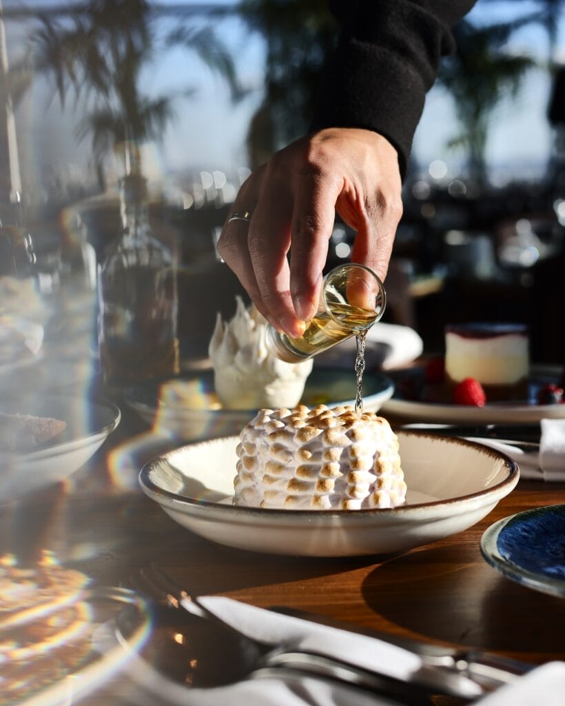 Une main, rappelant une scène de Lucy dans le ciel, verse un liquide sur un dessert à la surface texturée dans un bol sur une table en bois, entouré de plats et de verres, sur un fond flou.