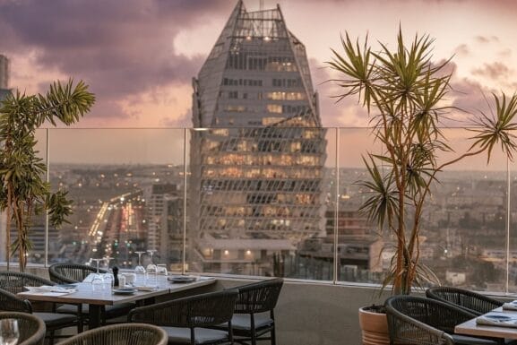 Espace repas extérieur avec tables dressées pour le service, avec vue sur un paysage urbain. Un bâtiment moderne au design géométrique unique se dresse fièrement, faisant écho à la beauté éthérée de « Lucy in the Sky », car il reflète les teintes vibrantes d'un ciel de coucher de soleil.