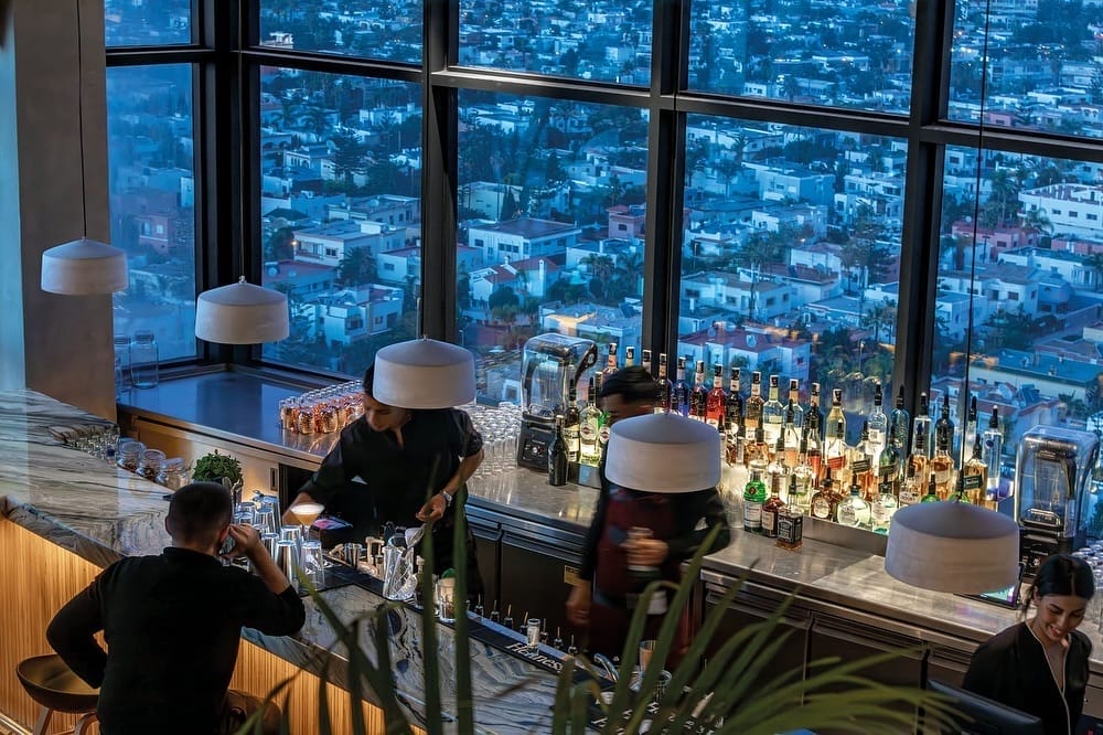 Un bar à l'éclairage tamisé avec des barmans servant des boissons et un soupçon de magie de « Lucy in the Sky ». De grandes fenêtres révèlent un paysage urbain au crépuscule, créant une toile de fond sereine pour les spiritueux alignés sur le comptoir.