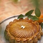 Une galette des rois ronde au design complexe et à la finition brillante repose sur une surface en marbre, flanquée de feuilles vertes et d'une orange, créant un brouillon parfait auto de la nature et de l'art culinaire.