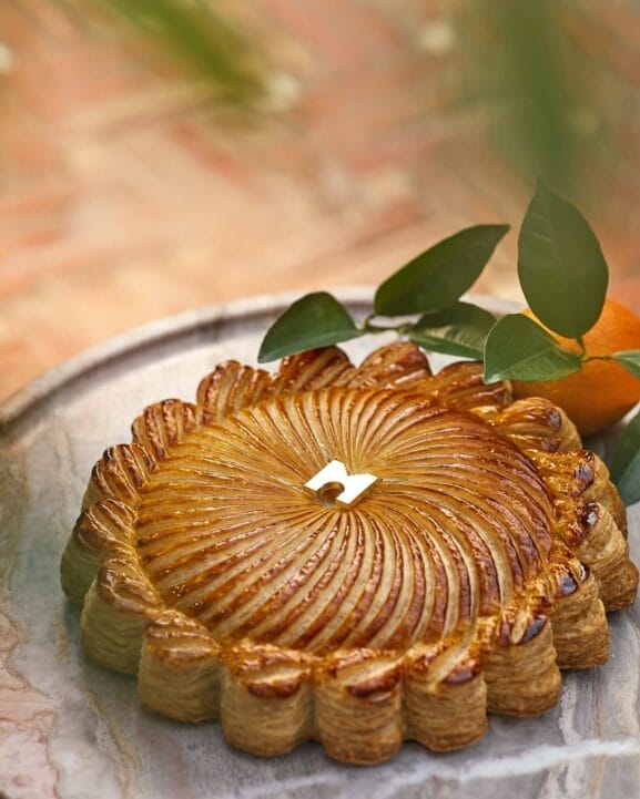 Une galette des rois ronde au design complexe et à la finition brillante repose sur une surface en marbre, flanquée de feuilles vertes et d'une orange, créant un brouillon parfait auto de la nature et de l'art culinaire.