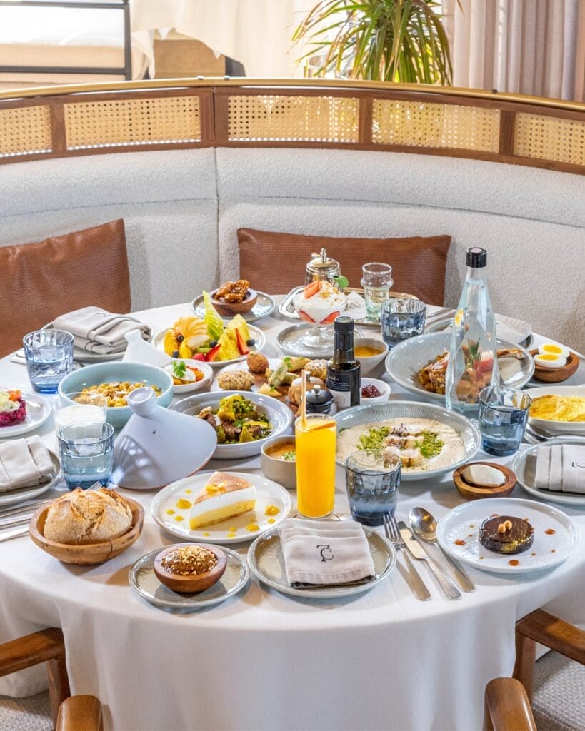 Une table ronde dressée avec une variété de plats de petit-déjeuner, de boissons et de pâtisseries sur une nappe blanche évoque l'essence du ftour à Casablanca, entourée de chaises en bois.