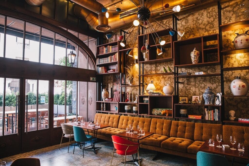 Intérieur de restaurant chaleureux avec un long canapé marron, des chaises colorées et des tables en bois dressées pour les repas. Des étagères décoratives accueillent des livres et des vases, tandis qu'une grande fenêtre offre une vue sur un patio extérieur parfait pour profiter du ftour à Casablanca.