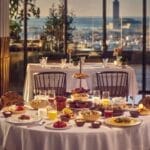 Une table à manger dressée avec une variété de plats et de boissons dans un restaurant avec de grandes fenêtres donnant sur le paysage urbain animé de Casablanca au coucher du soleil, créant un cadre idéal pour une expérience de ftour inoubliable.