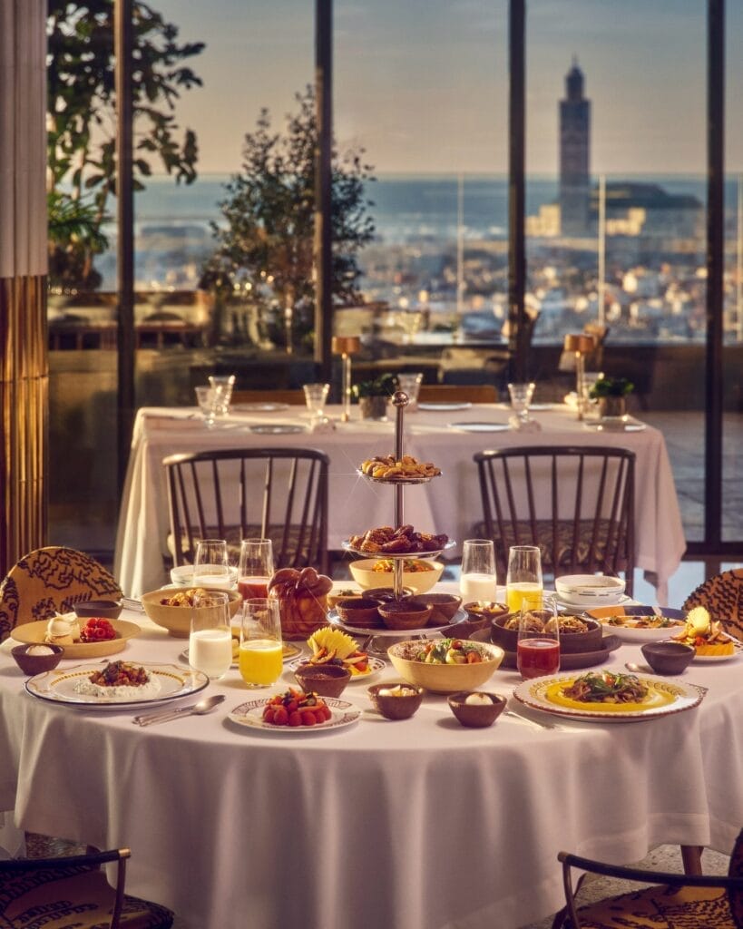 Une table à manger dressée avec une variété de plats et de boissons dans un restaurant avec de grandes fenêtres donnant sur le paysage urbain animé de Casablanca au coucher du soleil, créant un cadre idéal pour une expérience de ftour inoubliable.