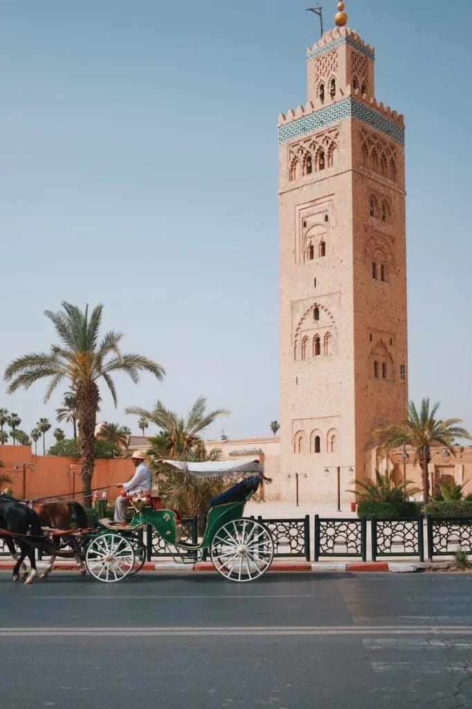 Une calèche passe gracieusement devant une haute tour ornée entourée de palmiers à Marrakech, sur fond de ciel bleu clair - une scène parfaite pour les activités familiales avec des enfants.