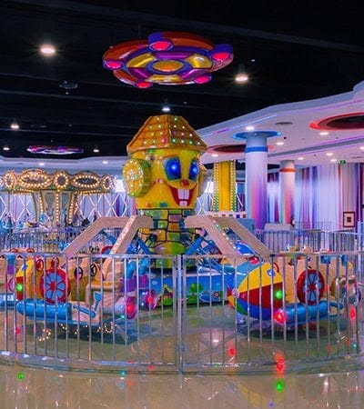 Manège d'intérieur coloré avec sièges sur le thème des vaisseaux spatiaux, idéal pour les activités familiales à Marrakech. Orné de lumières colorées et d'un chapiteau lumineux suspendu au plafond, il est entouré d'une clôture métallique qui assure la sécurité des enfants qui explorent le parc.