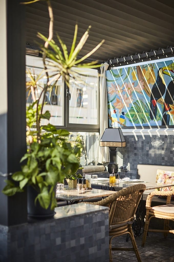 Un patio ensoleillé avec des chaises en osier, une plante en pot, une table dressée pour les boissons et une peinture murale colorée représentant des oiseaux et du feuillage tropical sur le mur apportent un charme automobile Brouillon vivant à votre espace extérieur.