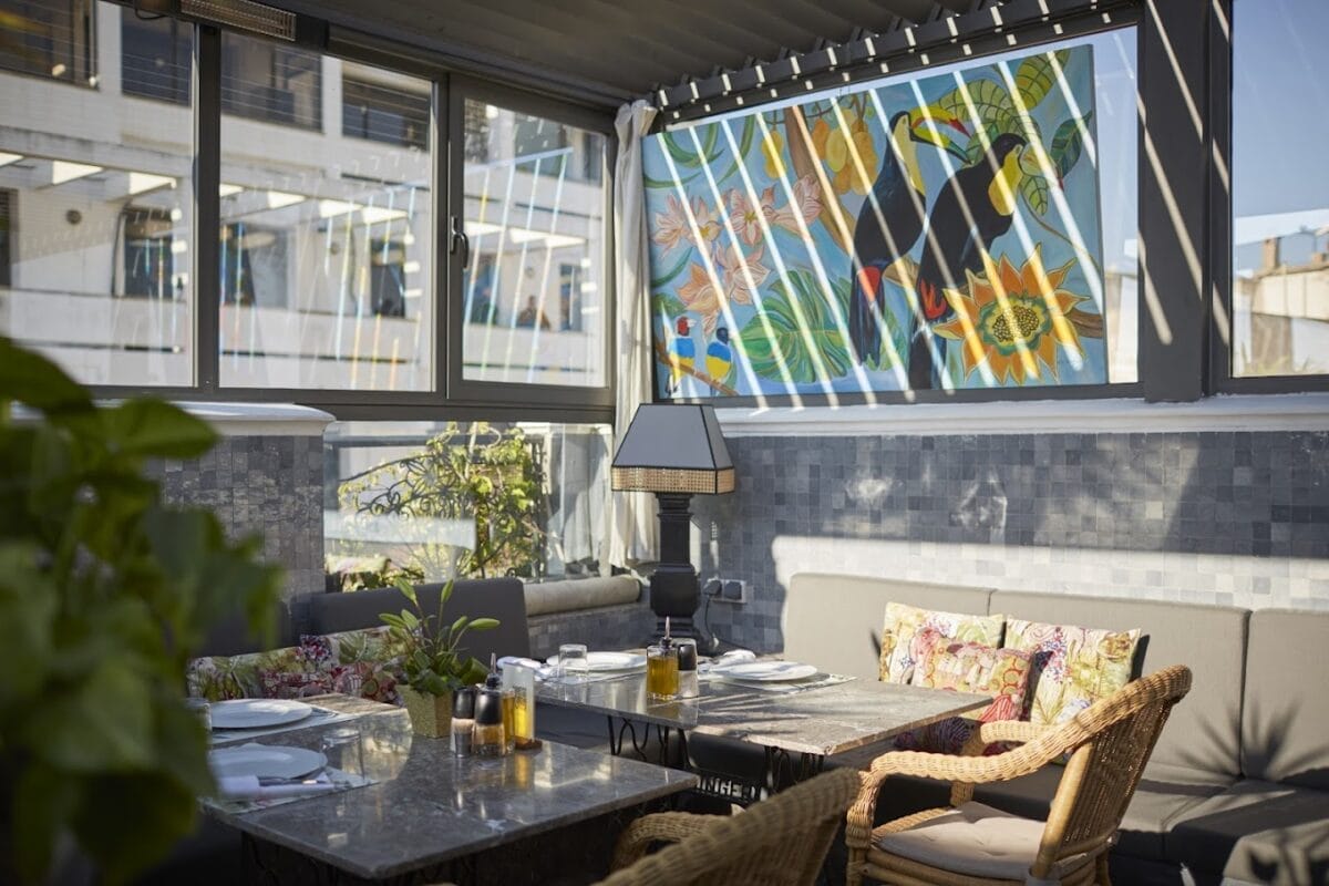 Une salle à manger intérieure ensoleillée avec une table en marbre, des chaises en osier, des plantes en pot et une peinture murale colorée représentant des toucans et des tournesols sur la fenêtre crée une ambiance automobile Brouillon détendue.