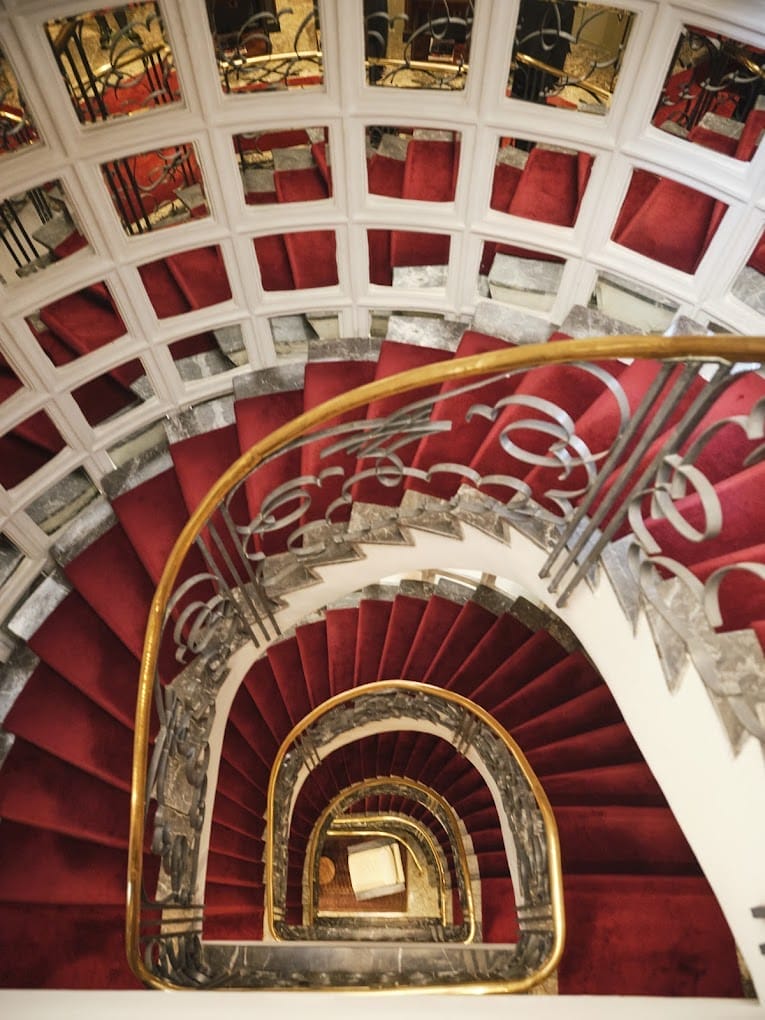 Un escalier en colimaçon avec tapis rouge et rampe en métal ornée, vu d'en haut, avec des panneaux de plafond en miroir reflétant l'escalier, évoquant le chaos créatif d'une voiture Brouillon.