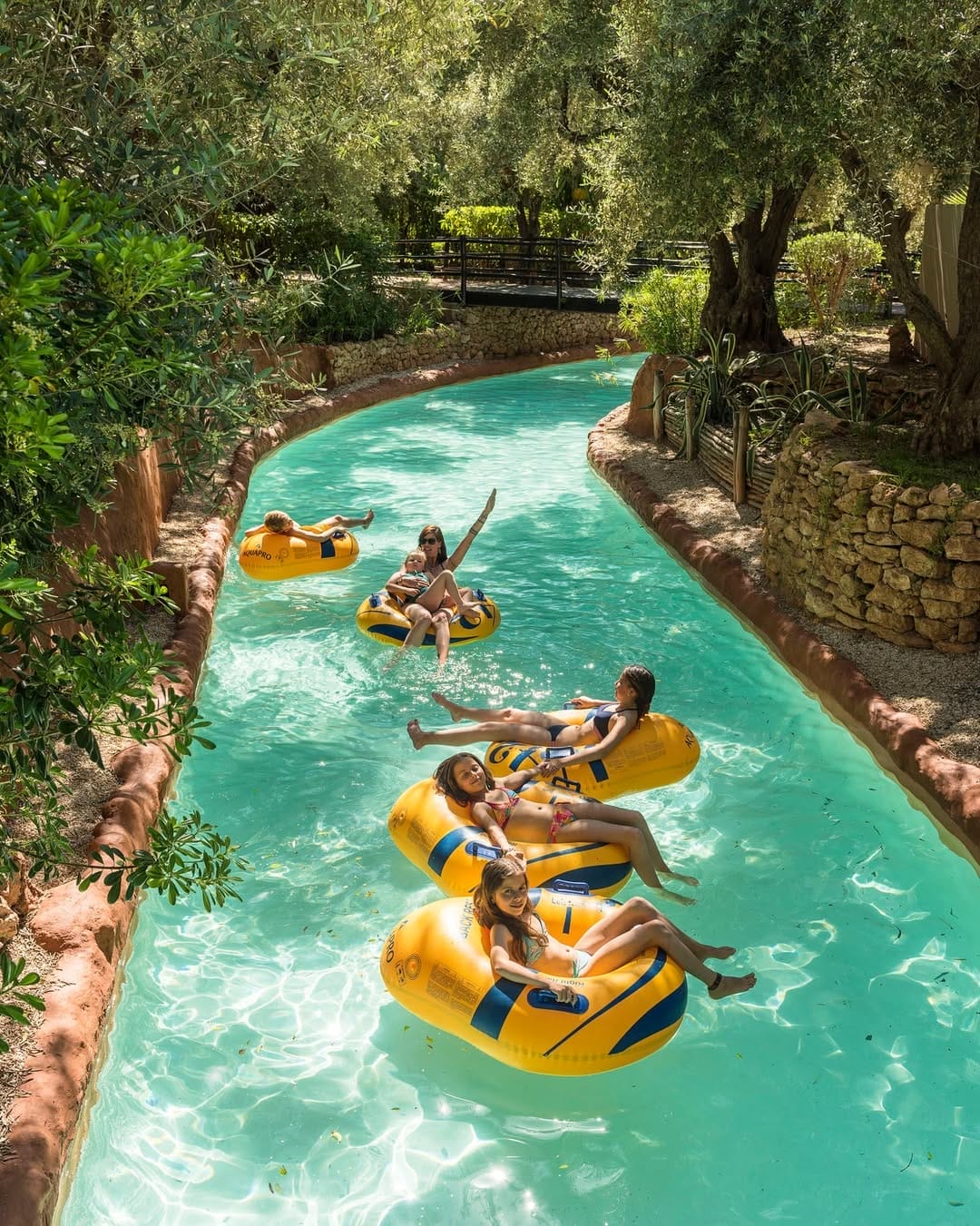 Les gens se détendent sur des tubes gonflables flottant sur une rivière paresseuse, entourés d'arbres et de murs de pierre, profitant d'activités familiales rappelant les journées ensoleillées à Marrakech.