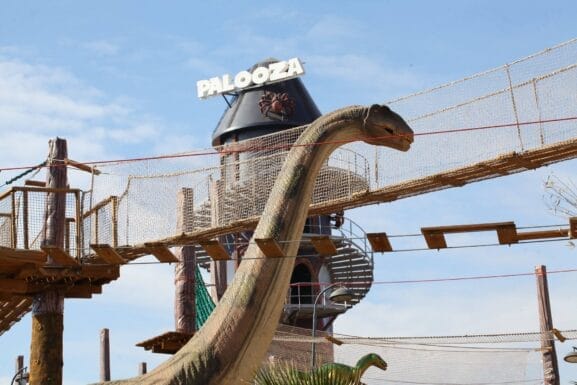 Une immense statue de dinosaure se dresse majestueusement sous un pont de singe au parc d'attractions, où le panneau « Palooza » se détache sur un ciel bleu azur. C'est l'endroit idéal pour des activités familiales, où parents et enfants profitent ensemble de cette aventure palpitante.