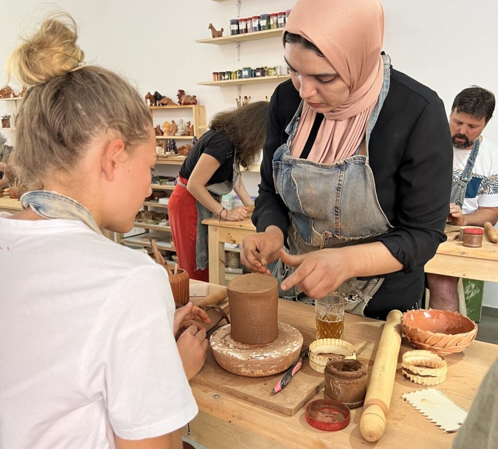Dans une salle de classe animée de Marrakech, une personne enseigne à une jeune fille l'art de la poterie. Entourée d'outils en argile et d'autres participants enthousiastes, cette activité est idéale pour les familles en quête d'expériences créatives avec leurs enfants.