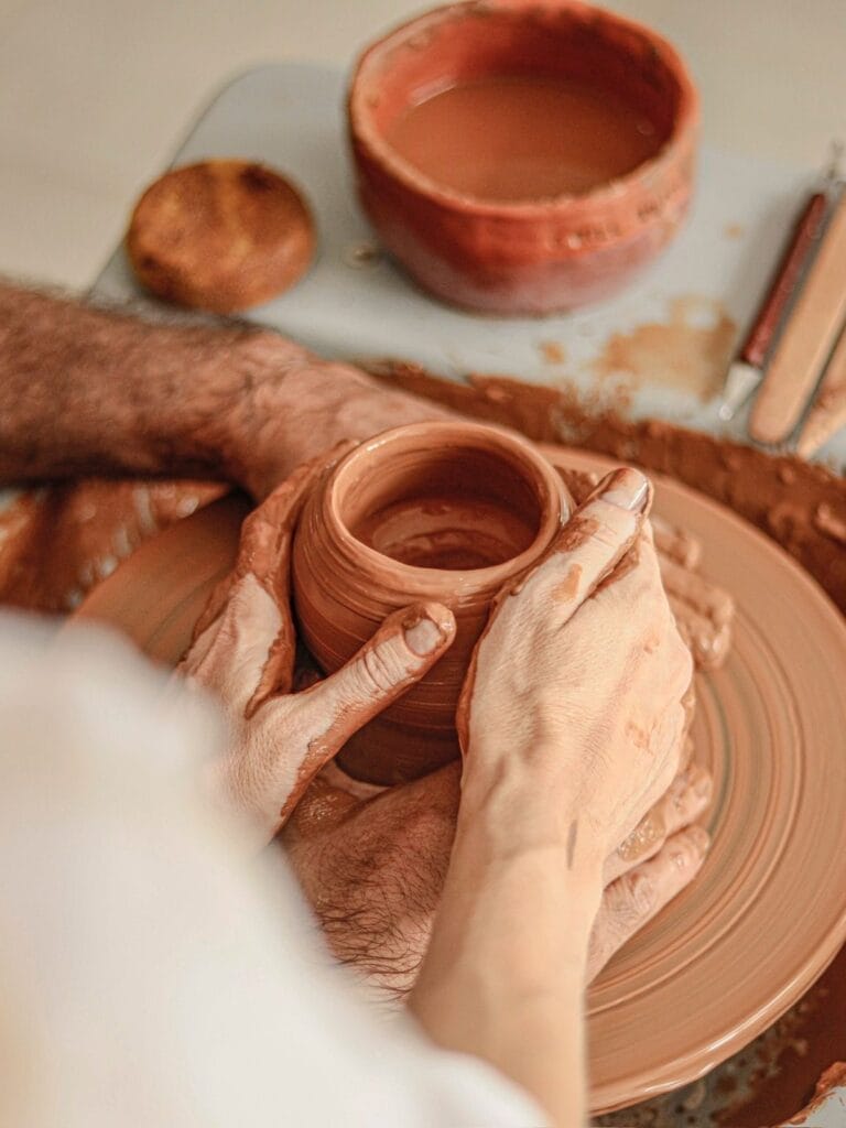 Deux personnes façonnent un pot en argile sur un tour de potier, leurs mains couvertes d'argile évoluant en harmonie. À proximité, un bol rempli de coulis et d'outils attend leur toucher. C'est un art à découvrir ensemble, une activité familiale intemporelle et appréciée de tous.