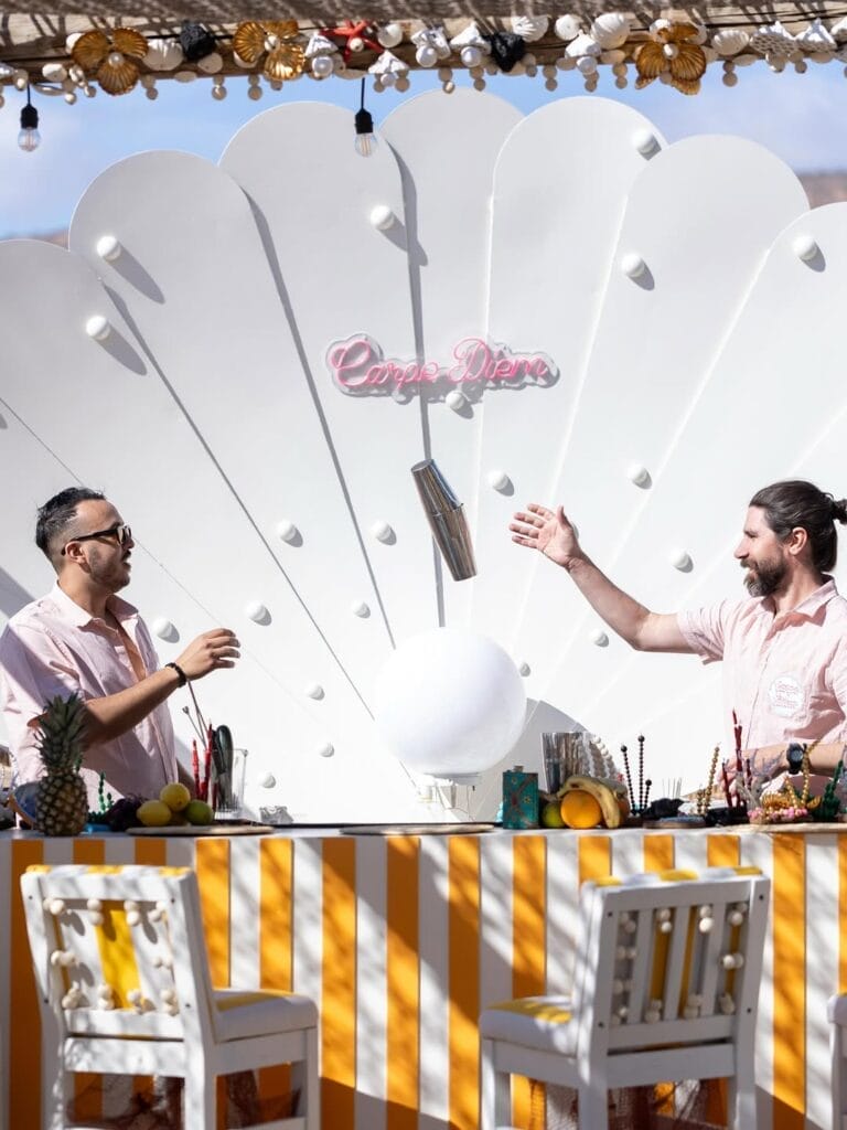 Deux hommes se tiennent derrière un bar rayé jaune et blanc avec des fruits et des boissons, servant des délices de l'été, devant une grande toile de fond en forme de coquillage avec une enseigne en néon "Carpe Diem".