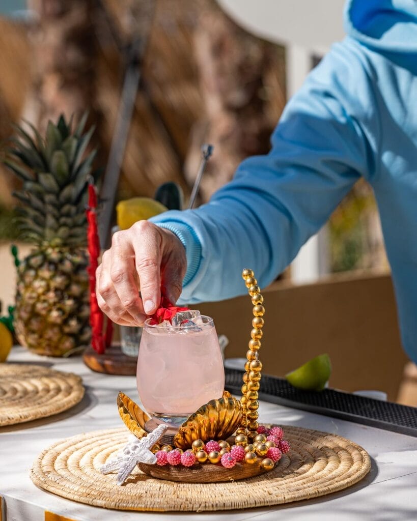 Une personne vêtue d'un sweat à capuche bleu garnit un cocktail rose dans un verre, posé sur un sous-verre doré orné de perles, sur un napperon tissé. L'ananas et le citron vert annoncent les délices de l'été à déguster à proximité.
