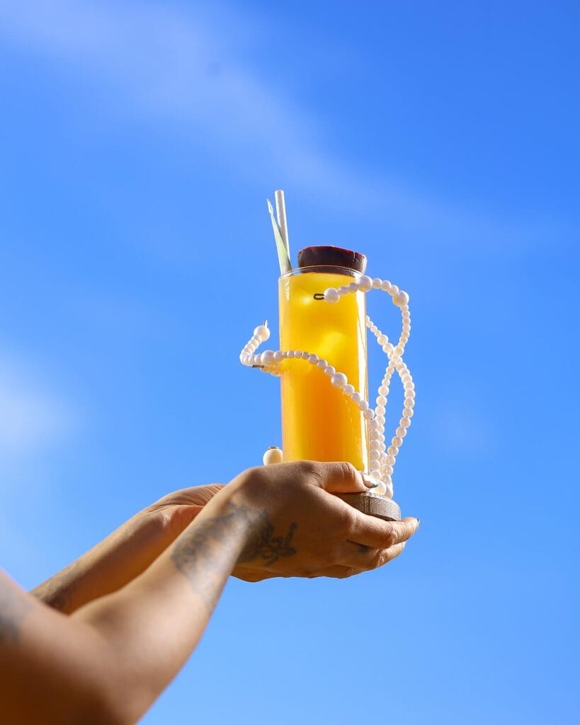 Une personne tatouée brandit un verre de jus d'orange orné de perles blanches et garni de feuilles, savourant les délices de l'été dans un ciel bleu limpide.