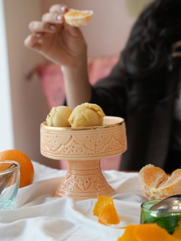 Une personne tient une tranche de mandarine épluchée au-dessus d'un plat décoratif contenant trois boules de glaces, avec des oranges et des pelures sur une table - parfait pour ceux qui cherchent des adresses branchées pour glaces Casablanca.