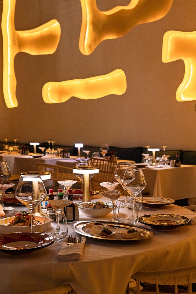 Une salle de restaurant avec des tables élégamment dressées, des éclairages décoratifs au mur, des verres à vin et des assiettes de délices de l'été inspirées de la côte atlantique.