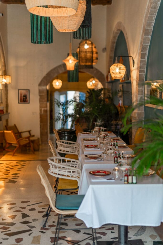 Un intérieur de restaurant avec des tables dressées, des nappes blanches, des chaises modernes, des lampes tressées suspendues et des plantes en pot le long des murs - parfait pour savourer les délices de l'été le long de la Côte Atlantique.