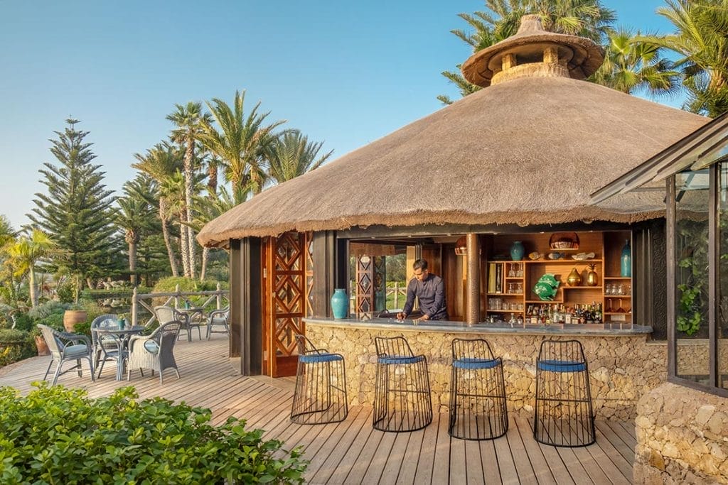 Bar en plein air avec toit de chaume, soubassement en pierre et terrasse en bois sur la côte atlantique, entouré de verdure et de palmiers ; un barman prépare des délices de l'été derrière le comptoir.