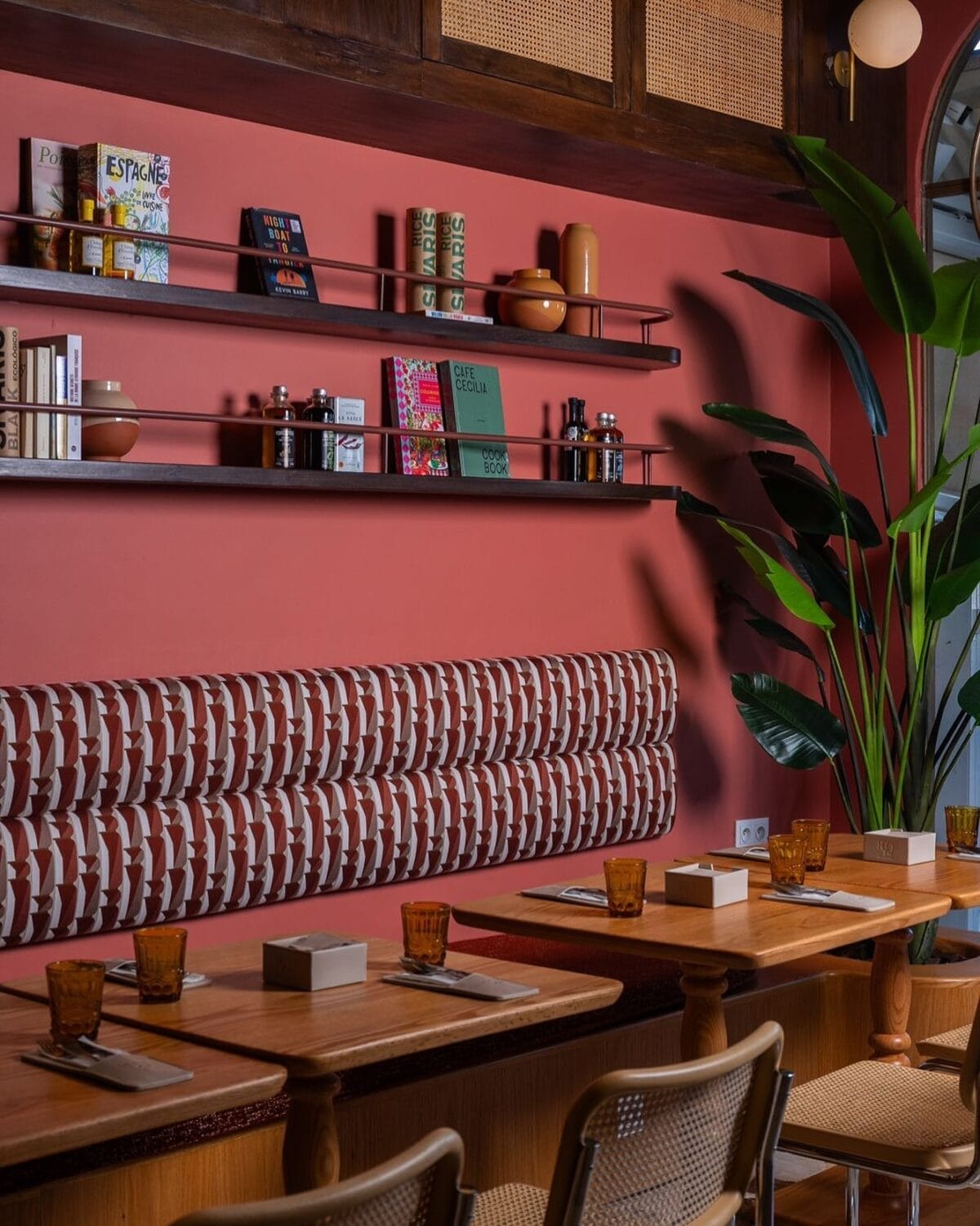 Intérieur d'un restaurant moderne avec des tables en bois, des banquettes à motifs, des étagères murales avec des livres et des objets de décoration, et une grande plante en pot.