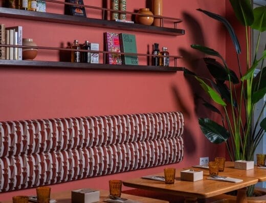 Intérieur d'un restaurant moderne avec des tables en bois, des banquettes à motifs, des étagères murales avec des livres et des objets de décoration, et une grande plante en pot.