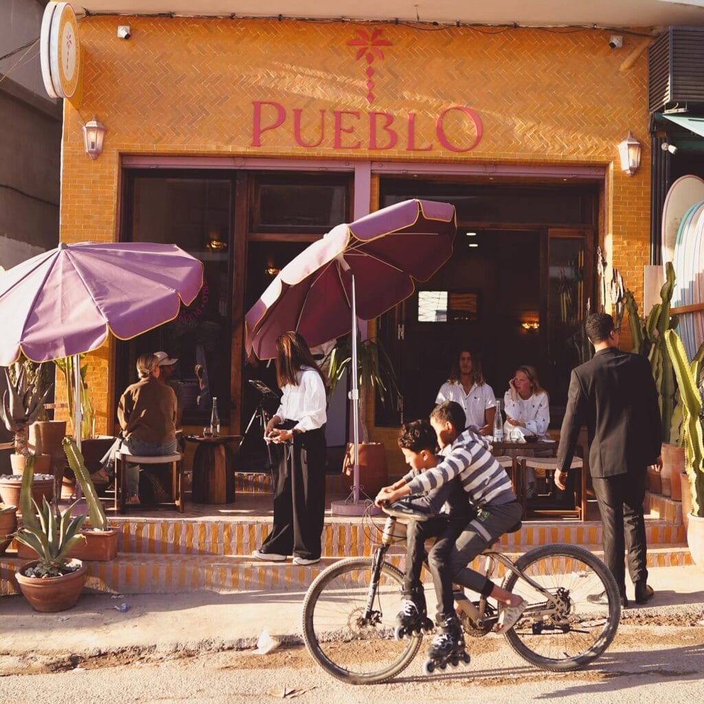 Des personnes sont assises et dînent à l'extérieur d'un restaurant appelé Pueblo, profitant d'une journée ensoleillée, tandis que deux garçons passent à bicyclette au premier plan, capturant ainsi un moment animé de l'automobile de Brouillon.