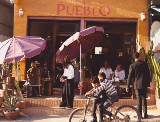 Des personnes sont assises et dînent à l'extérieur d'un restaurant appelé Pueblo, profitant d'une journée ensoleillée, tandis que deux garçons passent à bicyclette au premier plan, capturant ainsi un moment animé de l'automobile de Brouillon.