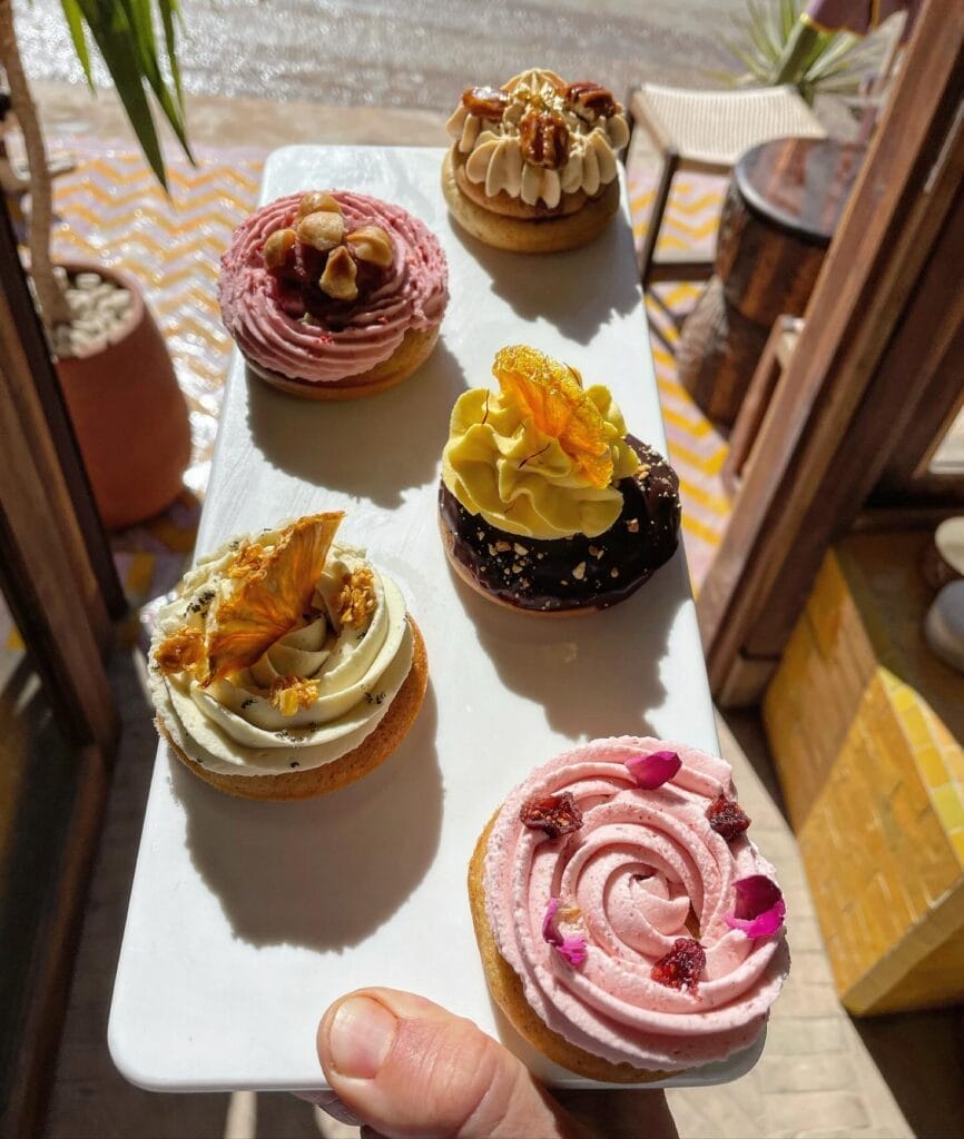 Une main tient un plateau blanc rectangulaire contenant six pâtisseries décorées, chacune garnie d'un glaçage coloré et d'un assortiment de garnitures, dans un décor intérieur ensoleillé - une scène invitante qui ressemble à un Brouillon auto de l'art du dessert.