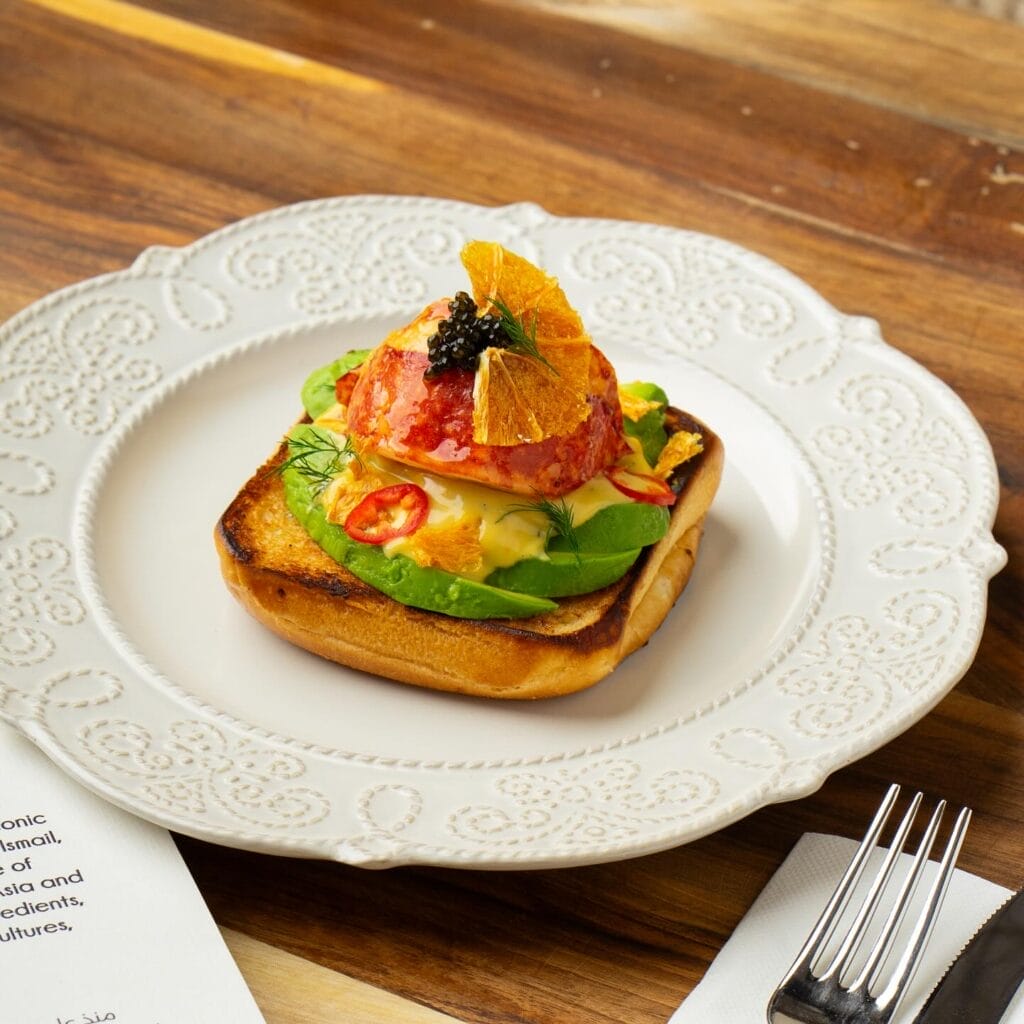 Un morceau de pain grillé garni de tranches d'avocat, de tomate, de piment, d'une cuillerée de sauce, de caviar et d'un croustillant à l'orange est servi avec art sur une assiette blanche - parfait pour être téléchargé en tant que Brouillon auto pour l'inspiration gastronomique.