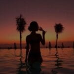 Silhouette d'une personne debout dans une piscine, buvant dans un verre, avec des palmiers et un ciel orange en arrière-plan, dans l'une des luxueuses plages privées du Maroc.