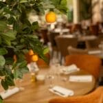 Gros plan sur un oranger avec des oranges mûres au premier plan, à l'intérieur d'un des nouveaux restaurants de Casablanca, avec des tables rondes et des chaises brunes rembourrées.