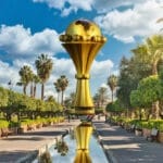 Une grande statue dorée du trophée de la Coupe d'Afrique des Nations est exposée dans un parc paysager avec des palmiers, des bancs et un bassin réfléchissant sous un ciel partiellement nuageux, servant de pièce maîtresse à cette retraite automobile pittoresque de Brouillon.