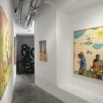 Un couloir de galerie d'art moderne - parfait pour les sorties culturelles à Casablanca - avec des peintures colorées encadrées sur des murs blancs, des sols en béton et des conduits apparents au plafond.